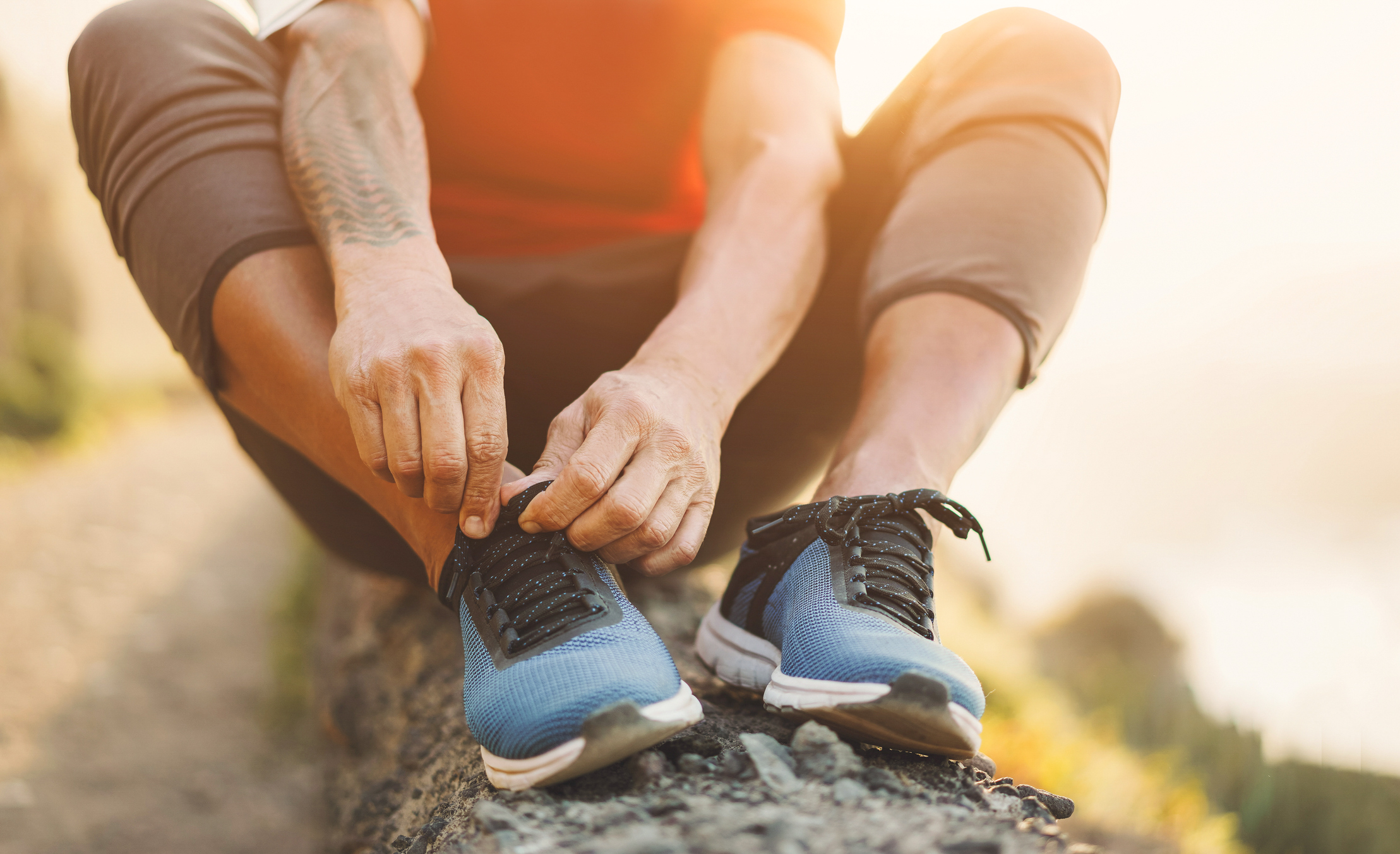 A person with medium skin tone, sitting on a fallen tree with athletic clothes on, tying a shoe.